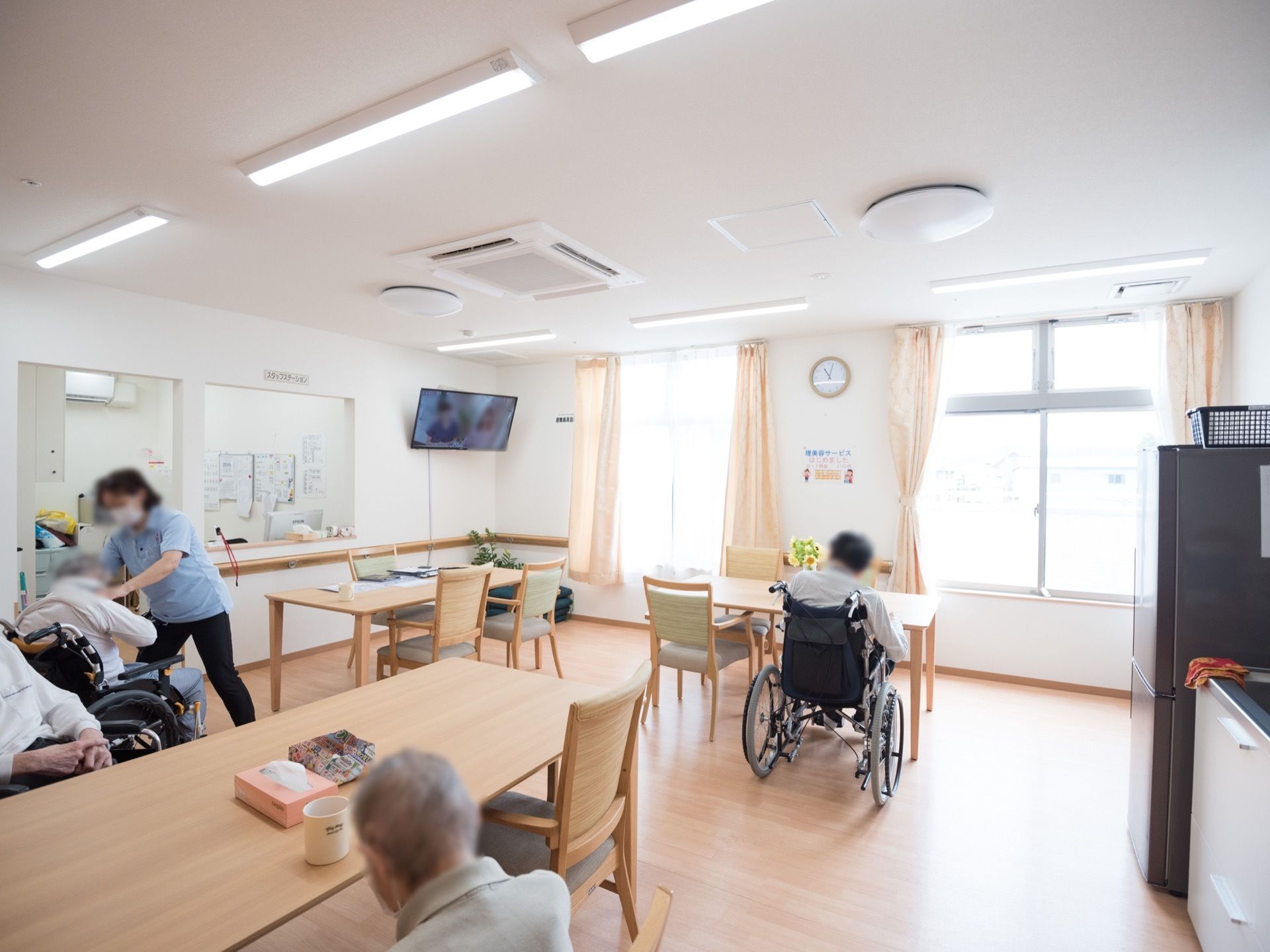 複数のテーブルや椅子が配置された明るい広間で過ごしている人々の姿が見える。右側に冷蔵庫や流し台がある。