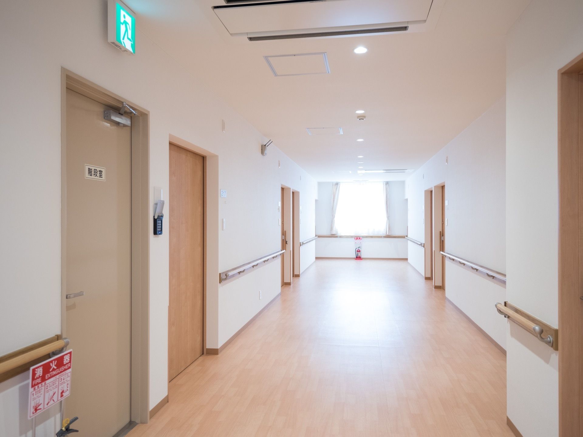 横幅の広い廊下の両側の壁面に、淡い色味の扉が設置されている。正面の壁面に窓が取り付けられており、その下方に消火器が置いてある。