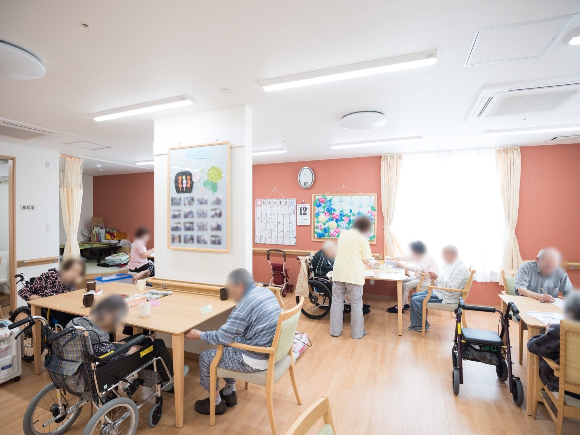 濃いピンク色の壁紙が施されたあたたかい雰囲気の広間にテーブルや椅子が並べられている。テーブルに向かいながら作業している人々の姿が見える。