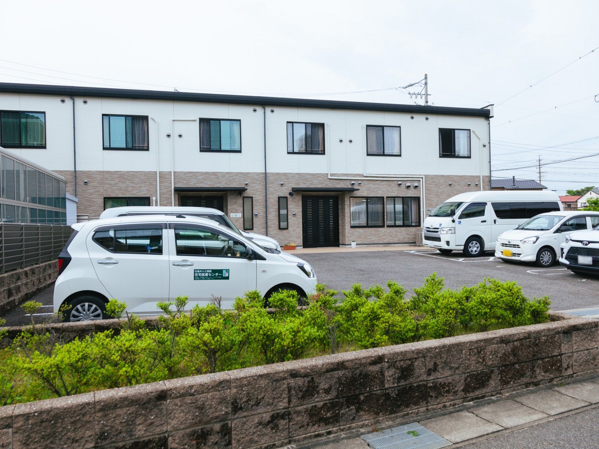 白とライトグレーの外壁の２階建ての建物は駐車場があり、草木が植えられている。