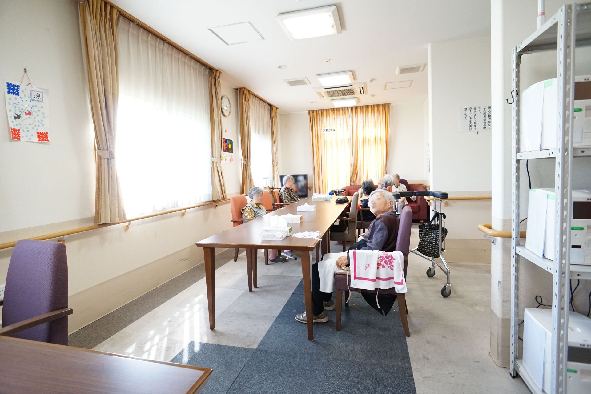 窓ガラスに囲まれた木製の机の周りに座る入居者様がいる広間
