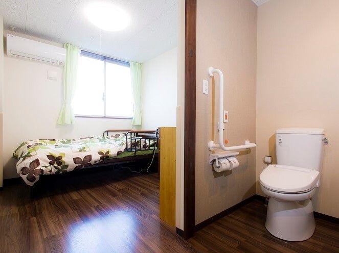 エル字型の手すりやオレンジの呼び出しボタンが付いたトイレはドアがなく、奥には寝具付きのベッドがある。