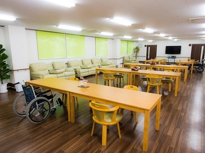 車いすや椅子がセットされた木製のダイニングテーブルが複数置かれたリビングダイニングにはソファもある。