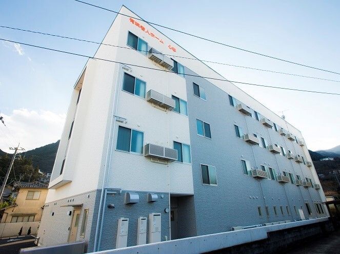４階建ての建物の周りには自然が多く、住宅もある。白とグレーの外観で、窓もたくさん付き、オレンジでホーム名が書かれている。