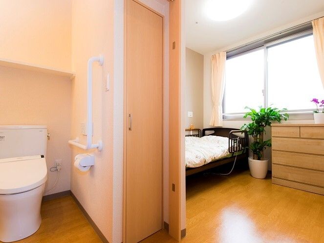 寝具付きのベッドが窓際にあり、観葉植物や花が飾られ、トイレにはエル字型の手すりやオレンジの呼び出しボタンがある。