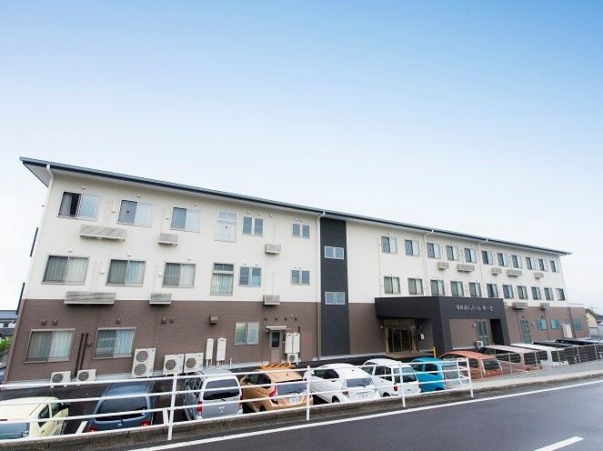 ３階建ての建物は白とブラウンで屋根が付いていて、玄関には車寄せが付いている。広い駐車場があり、たくさんの車が並んでいる。