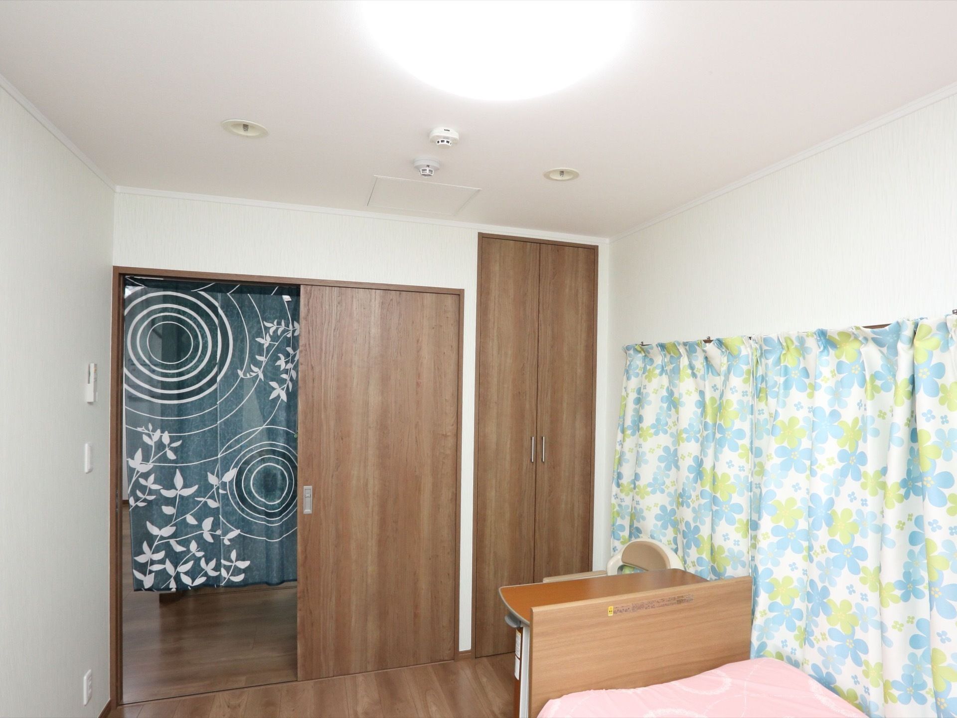 出入口のドアは開いており、外側にのれんが掛けられている。ベッドの側の窓には、花柄のカーテンが引かれている。