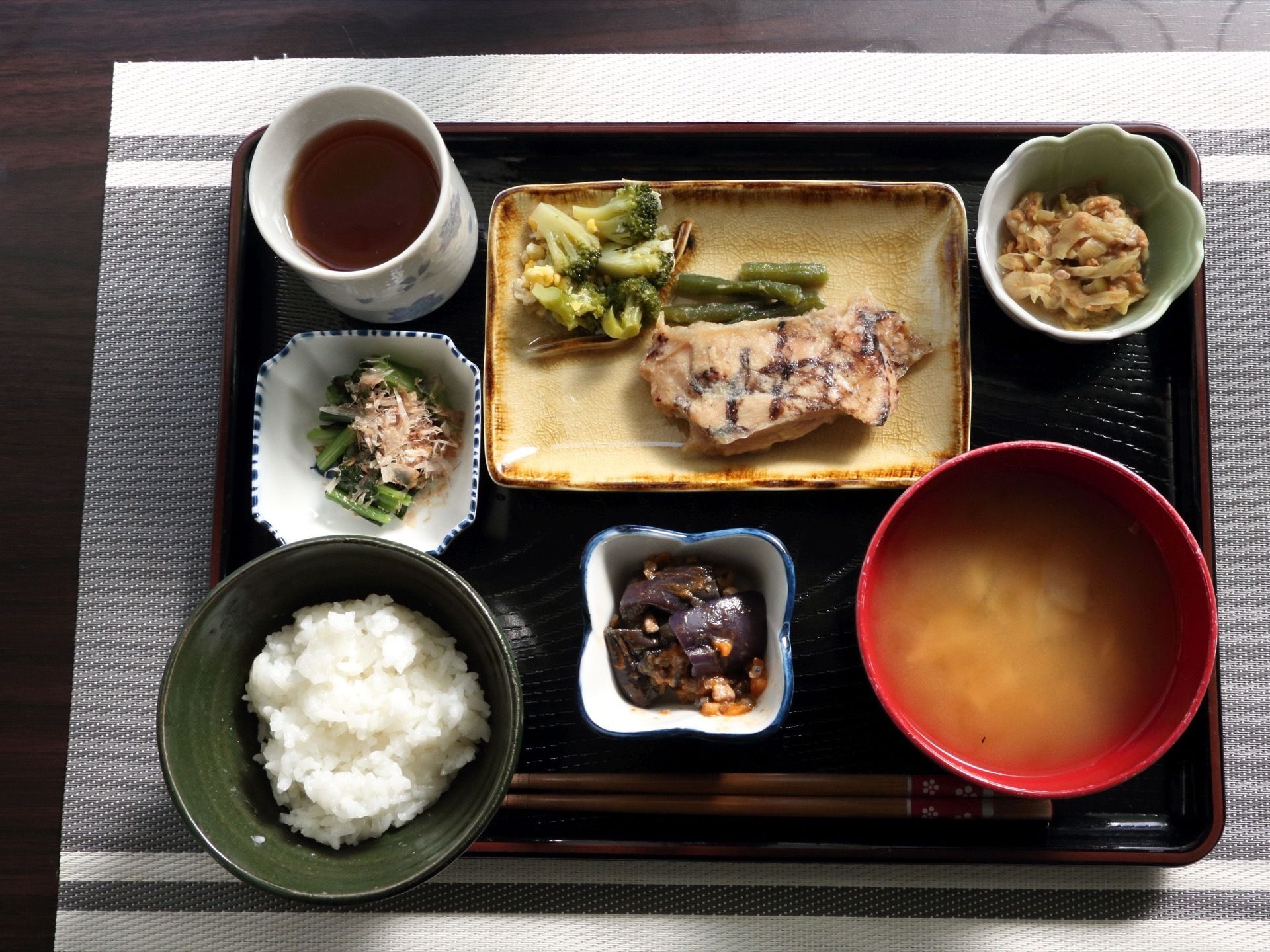 黒いトレーの上に食事が並べられている。ご飯、みそ汁、煮物、おひたしなどが用意され、お箸とお茶も添えられている。