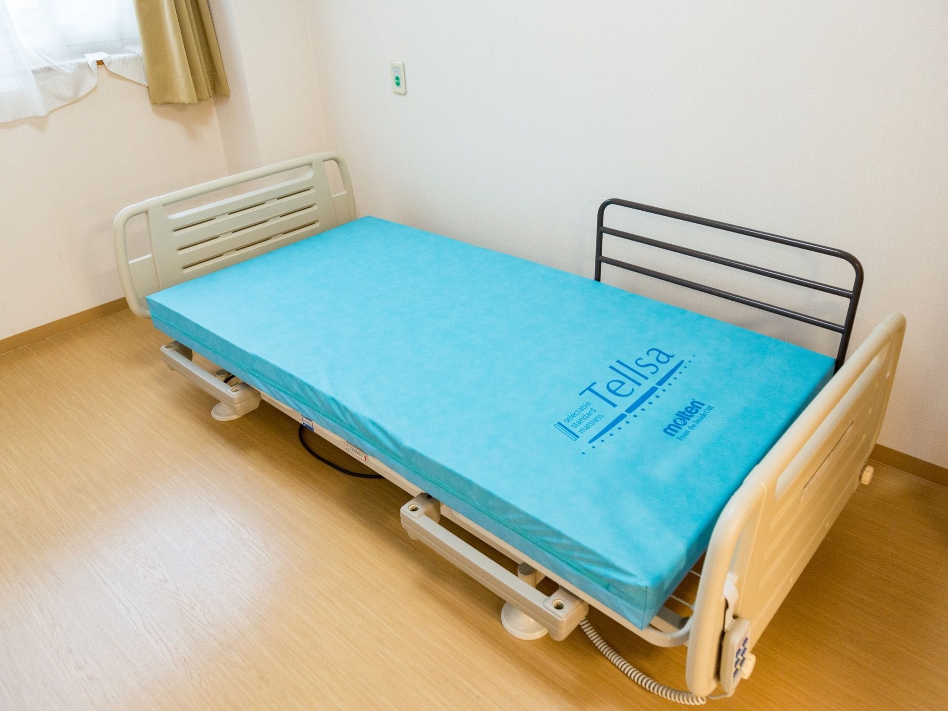 居室内には、分厚いマットレスの敷かれたシングルベッドが置かれている。奥には、ベージュ色のカーテンが掛けられた窓も設置されている。