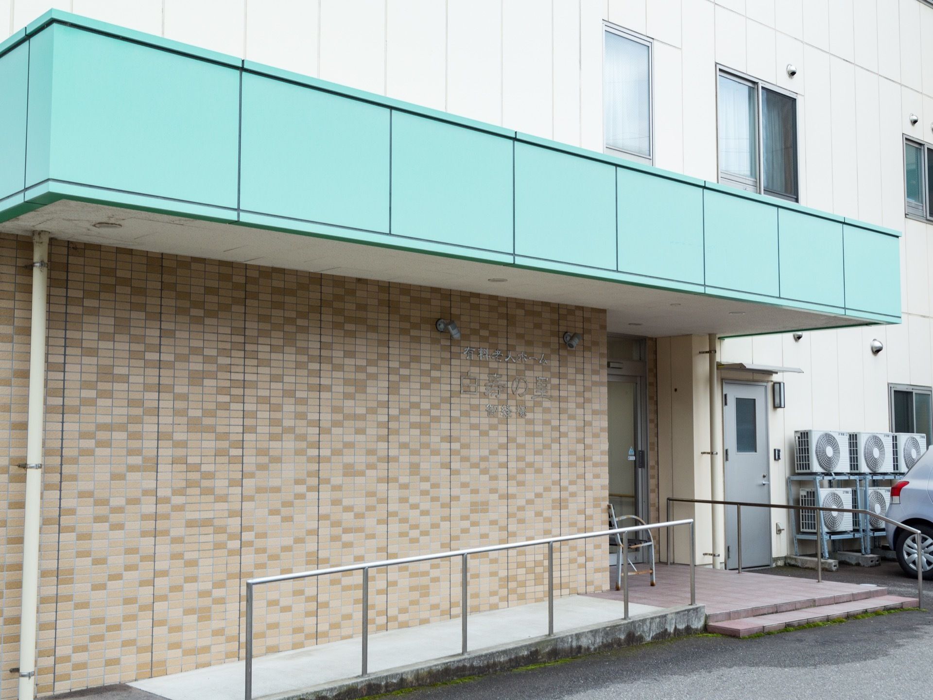玄関前の壁はレンガ調で、自動ドアが設置されている。スロープの両サイドには、シルバーの手すりが取りつけられている。