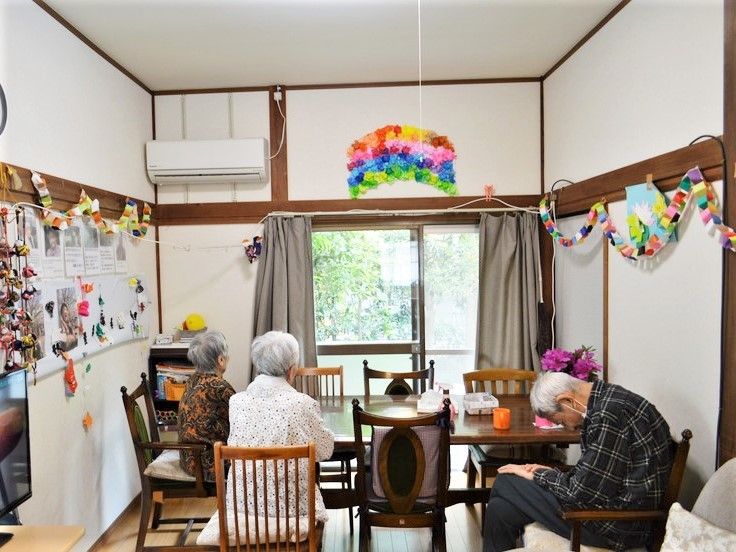 大きな木製のダイニングテーブルが中央に置かれ、入居者がテレビを見たりくつろいだりしている様子。