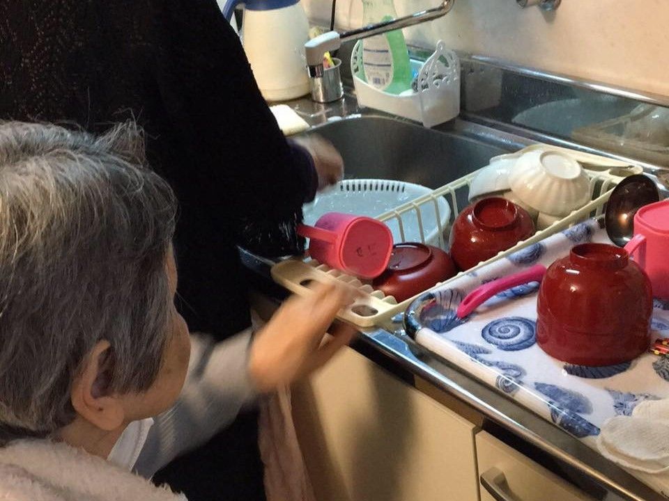 家庭的な雰囲気のキッチンで皿洗いが終わった食器をトレーに並べる入居者。