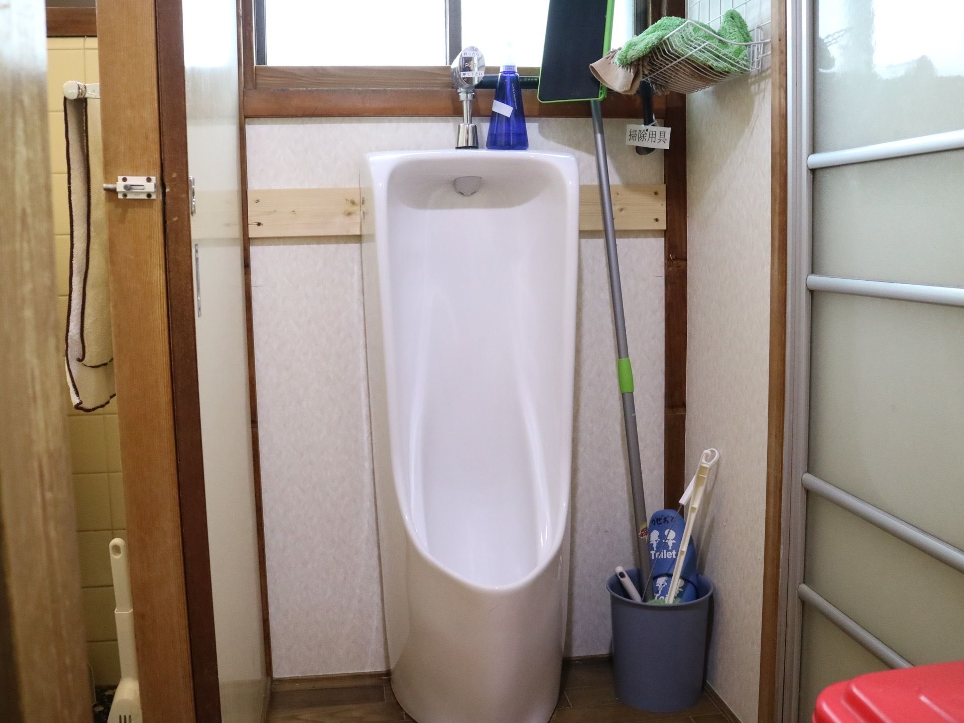 男性用のトイレである。足元には掃除用具がまとめられて置かれている。目の前には窓がある。床はフローリングである。
