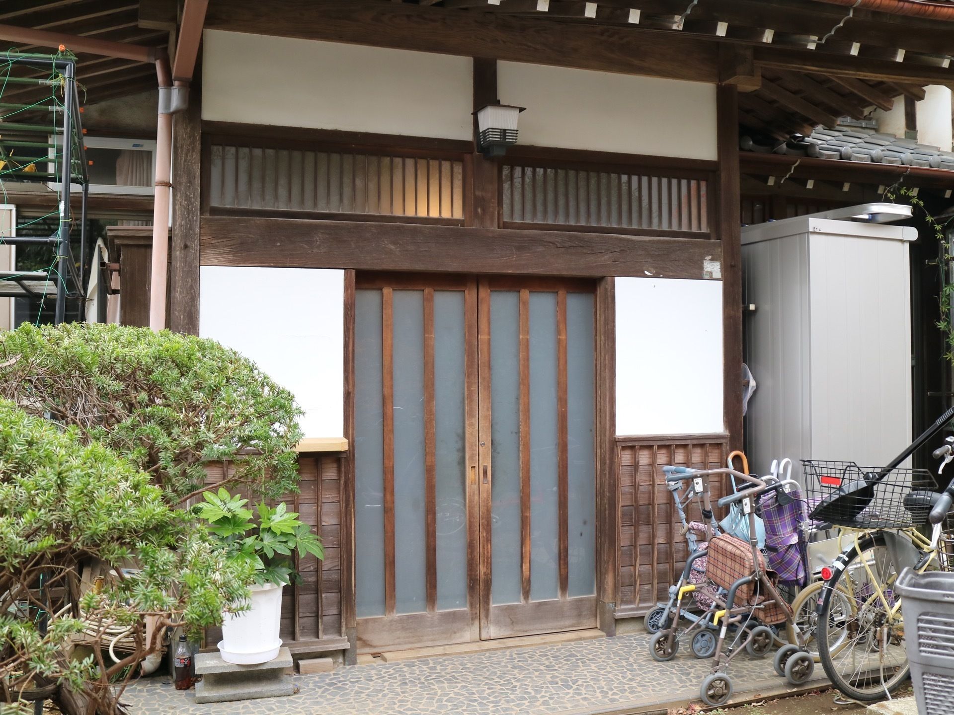 昔ながらの家である。自転車、シルバーカーが置かれている。奥には物置がある。プランターには植物が育てられている。
