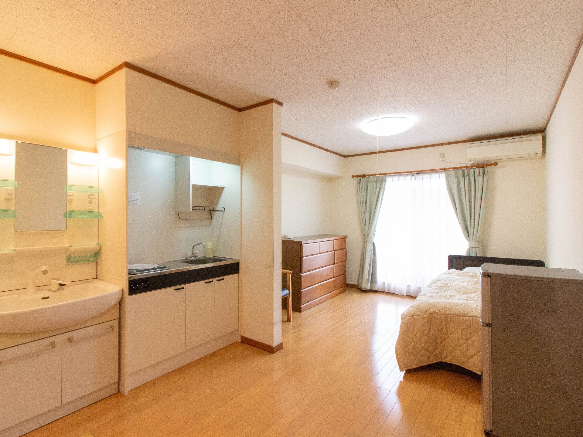 キッチンやベッド、洗面所や冷蔵庫などがついた明るいフローリングの居室。