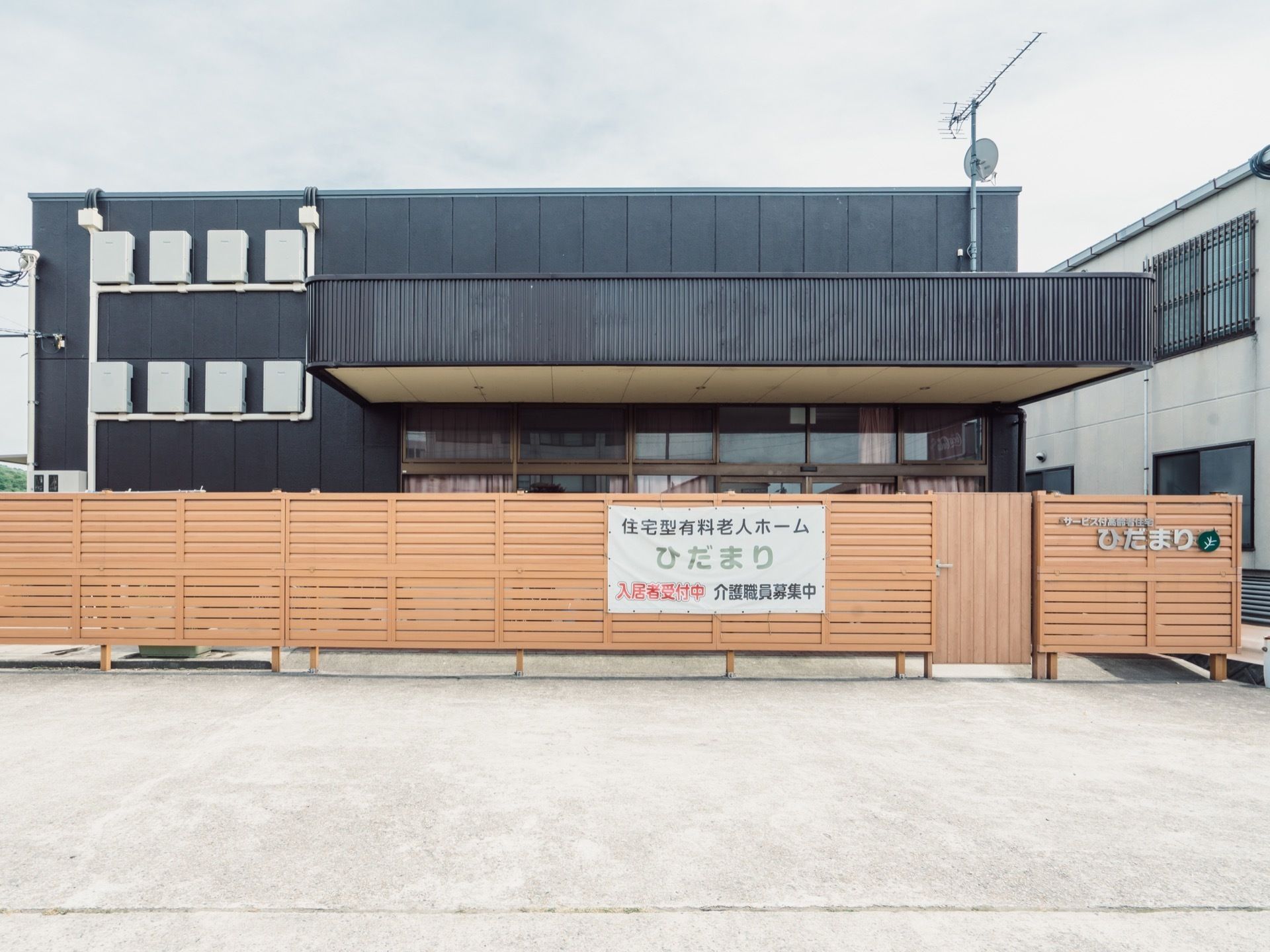 黒い外装の建物を覆うように木製の塀が設置されている。塀には施設名の銘板や募集ののぼりが取りつけてある。