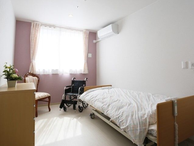 エアコンが付いた部屋には寝具がセットされたベッドやテーブル、タンスがあり、ベッドのそばには車いすも置かれている。