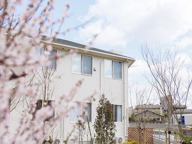 桜の花が咲いた枝が手前にあり、奥には2階建ての建物が建っている。建物の周りにはラティスも設置されている。