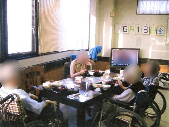 入居者たちが食事を摂っている。車椅子の入居者が３名、イスに座った入居者が１名いる。