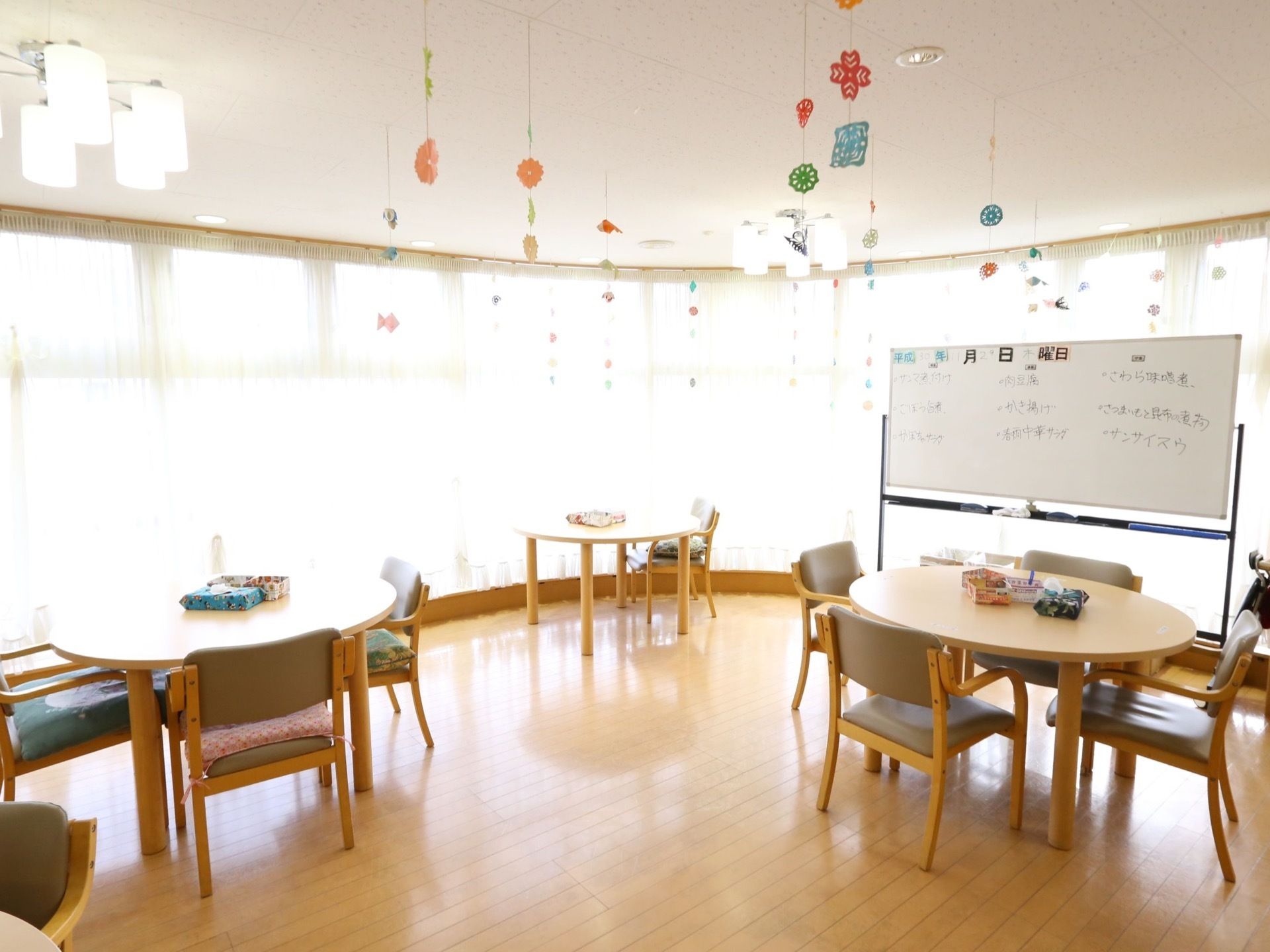 丸いテーブルが３つあり、その周りに椅子も複数置かれている。また、ホワイトボードには、食事のメニューが書かれている。