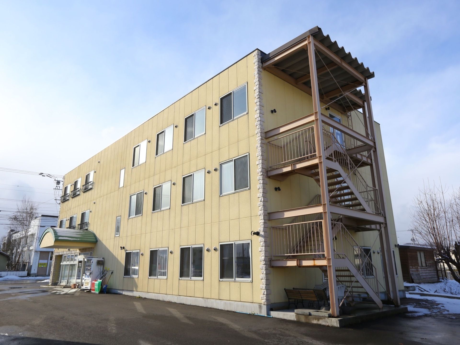 外階段のある、３階建ての建物である。周囲には雪が残っており、空は青空である。玄関横には、雪かき用の道具が置かれている。