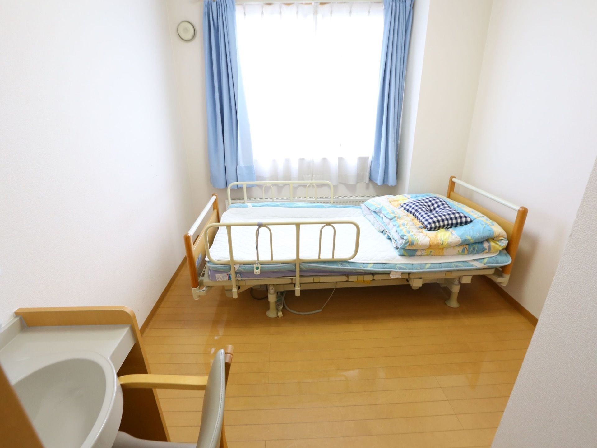 木目調の床に白い壁の居室には、水色のカーテンが掛かる窓と、洗面スペースがある。布団を敷いたベッドが置かれている。