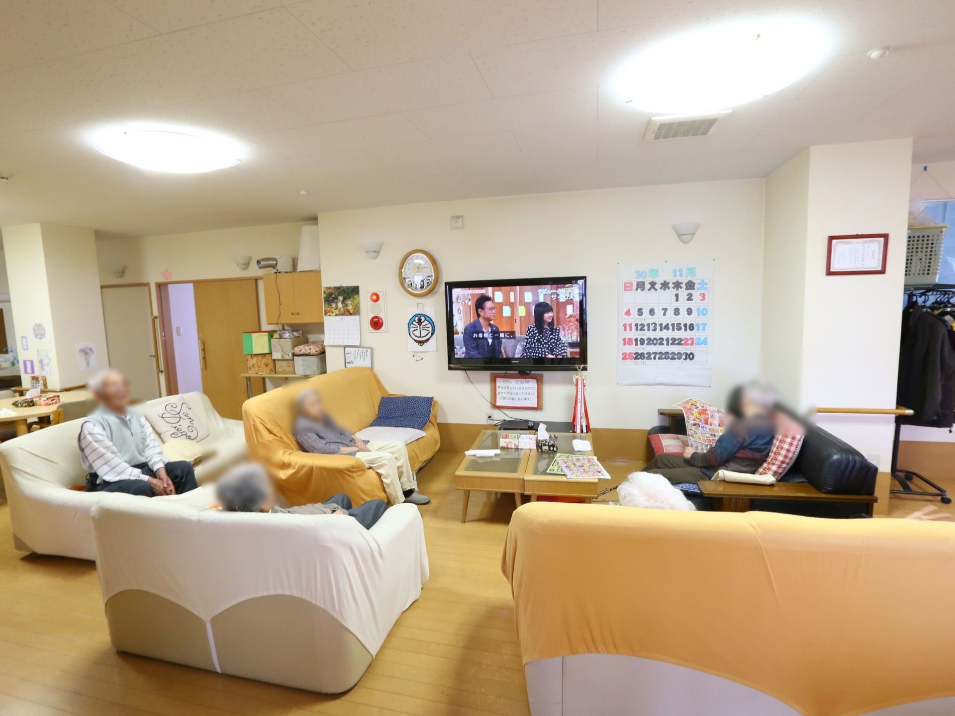 壁掛けのテレビとソファが5つ置かれたリビングである。ソファに座った入居者様がテレビを楽しんでいる。壁には大きなカレンダーが掛かっている。