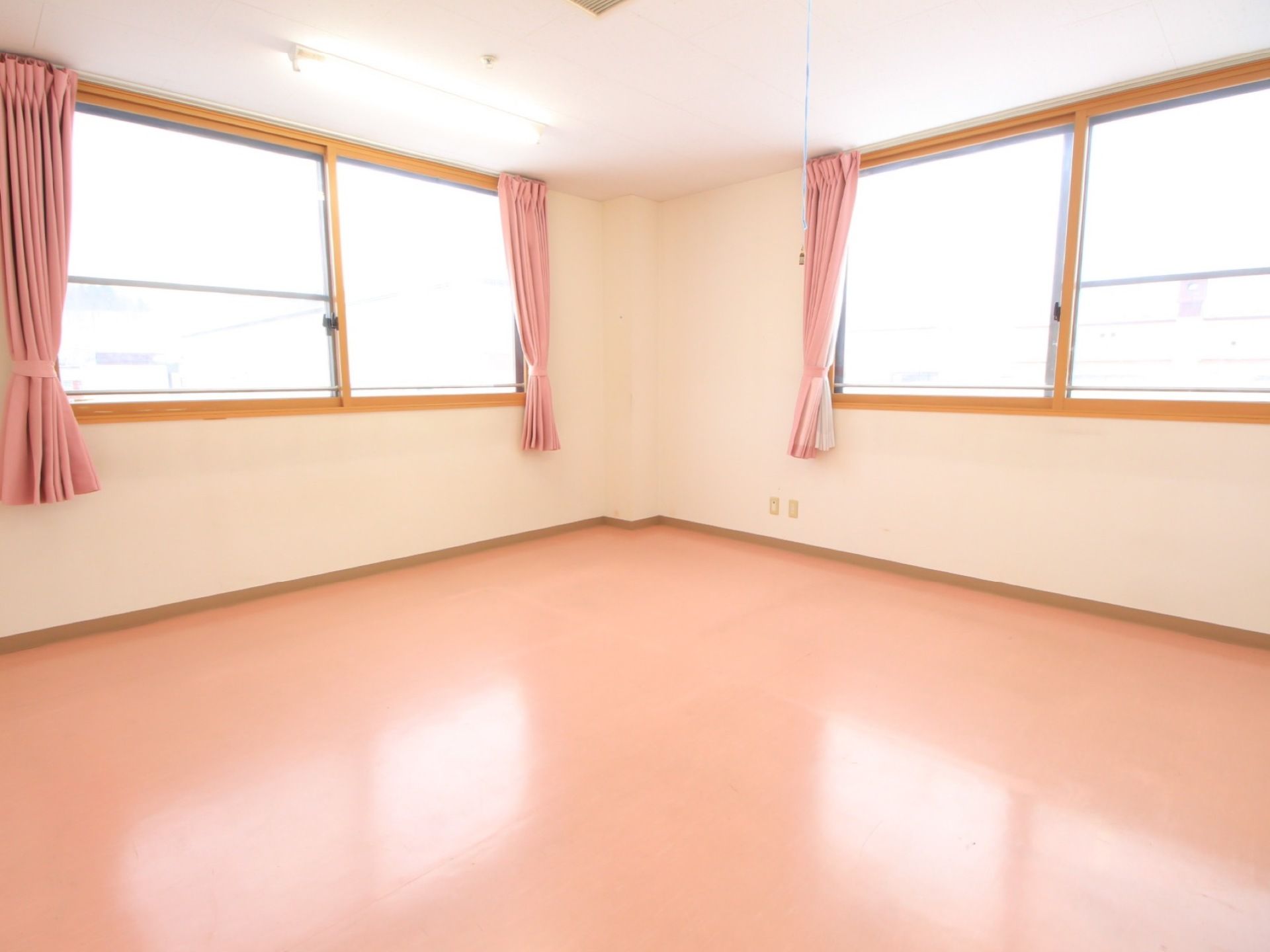 大きな窓が壁の２面に設置されたゆとりある居室である。床とカーテンがピンク色になっており、明るい雰囲気である。