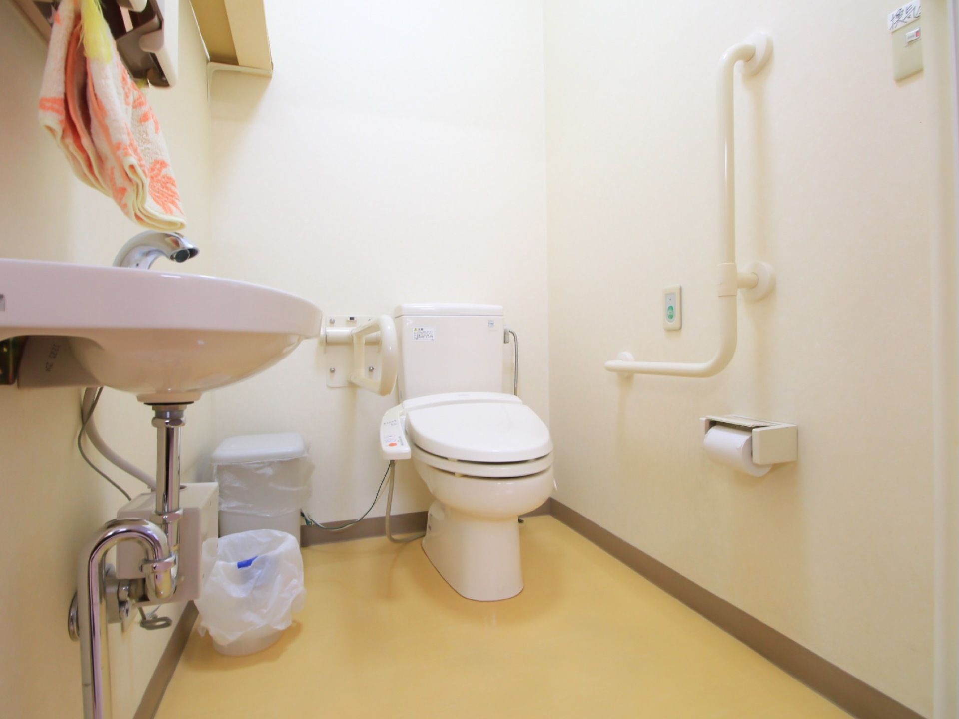 白い壁で清潔感のあるトイレである。壁にＬ字型の手すりが設置されており、温水洗浄便座が採用されている。