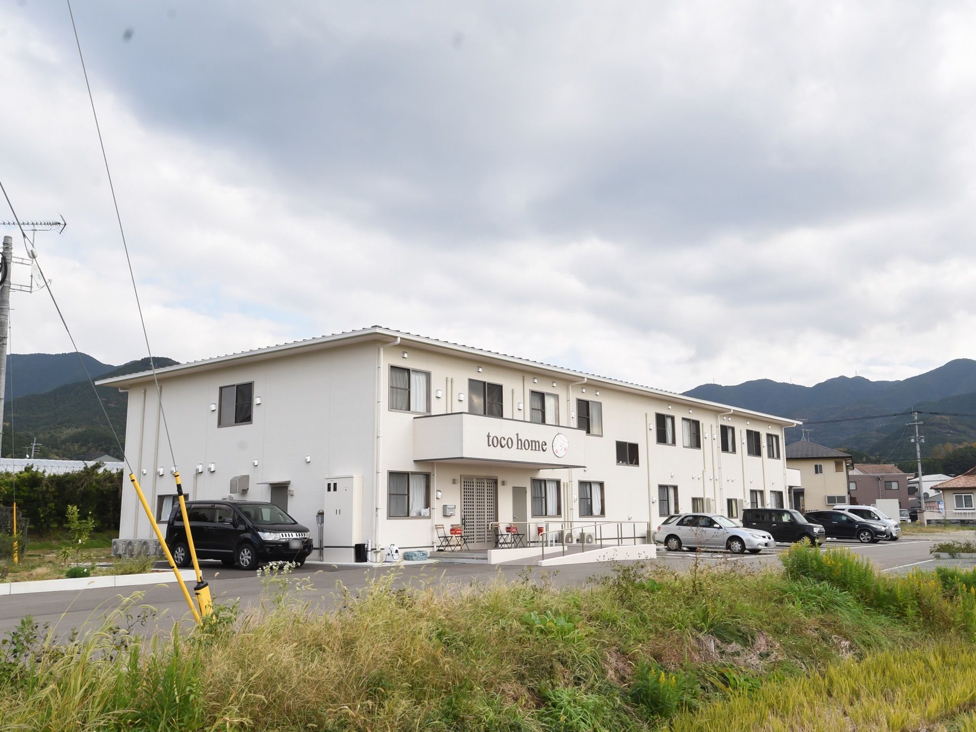 施設は緑や田んぼが広がる立地にある。周辺に高い建物がなく開放的である。駐車スペースを広く取っている。