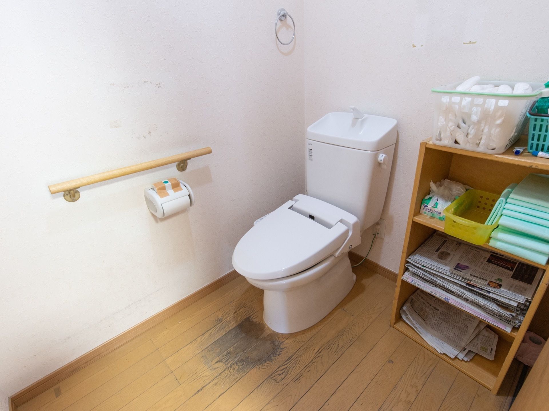 トイレの壁に手すりがあり、その下にトイレットペーパーホルダーが設置されている。便座の横には収納ラックが置かれている。