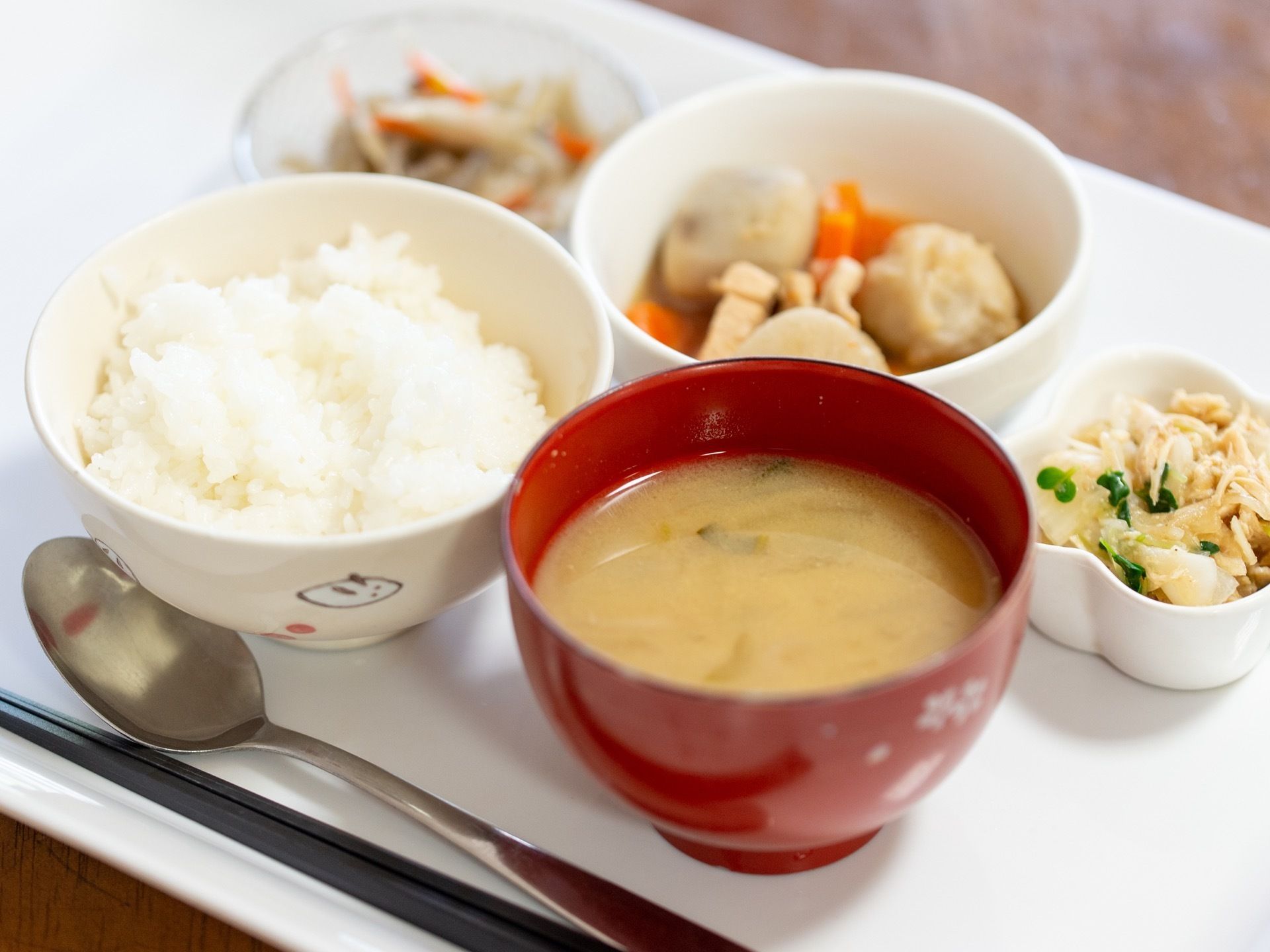 白いプレートの上にご飯、味噌汁、煮物、サラダの小鉢などが並べられており、スプーンと箸が用意されている。