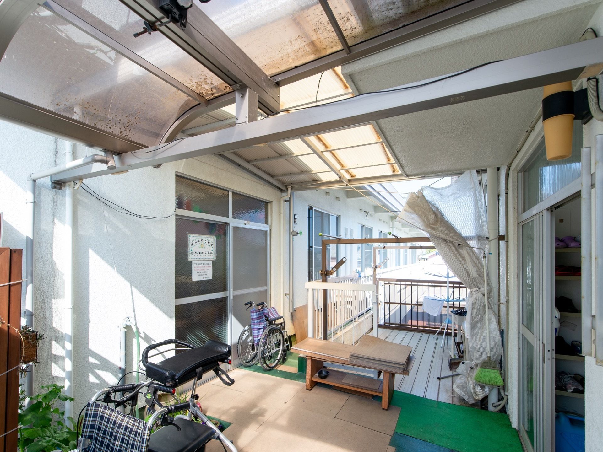 半透明な素材の屋根を掛けられた空間である。折りたたみ車椅子が2台置かれており、建物の中に下駄箱が見える。
