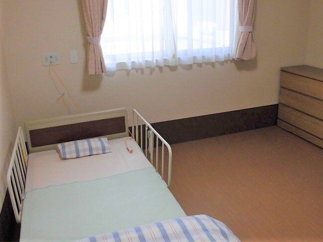 寝具がセットされた介護ベッドが置かれた居室には大きな窓があり、カーテンとレースカーテンが付き、木製のタンスも置かれている。
