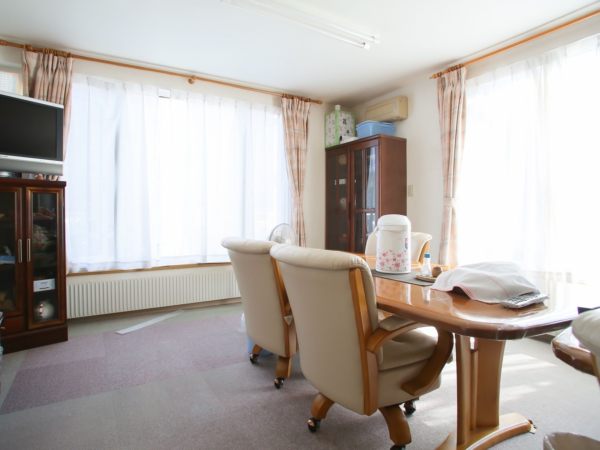 テーブルの真ん中に電気ポットや茶わんなどが置かれている。部屋の角にある棚やタンスは光沢のあるダークな材質となっている。