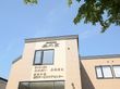 建物はベージュ系の外壁になっており、上の方に施設名が書かれている。窓やライトが付いていて、近くには木が植えられている。