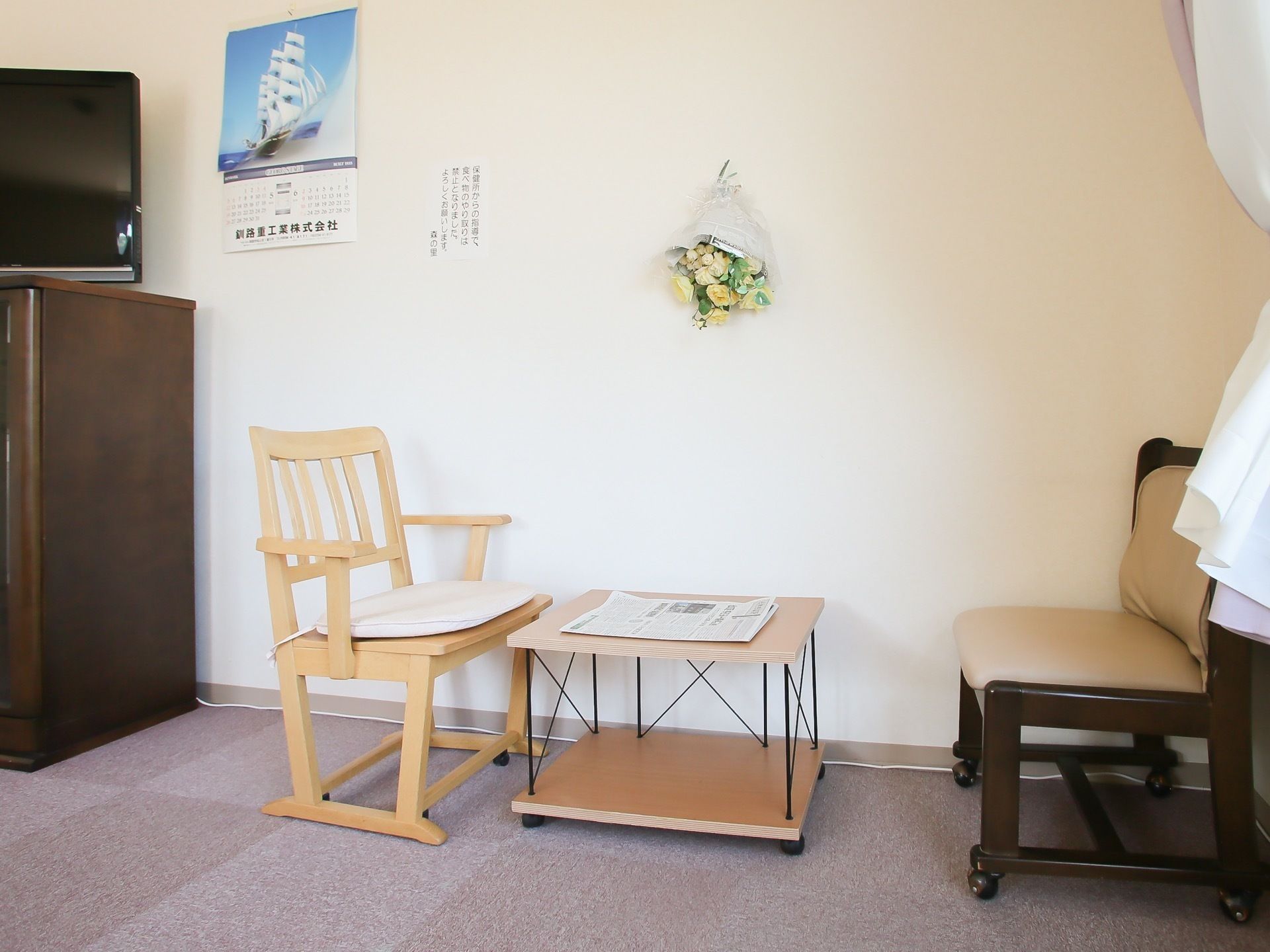 小さな木製テーブルに向き合って、椅子が２脚セットされている。後ろの壁には、ドライフラワーとカレンダーが掛けられている。