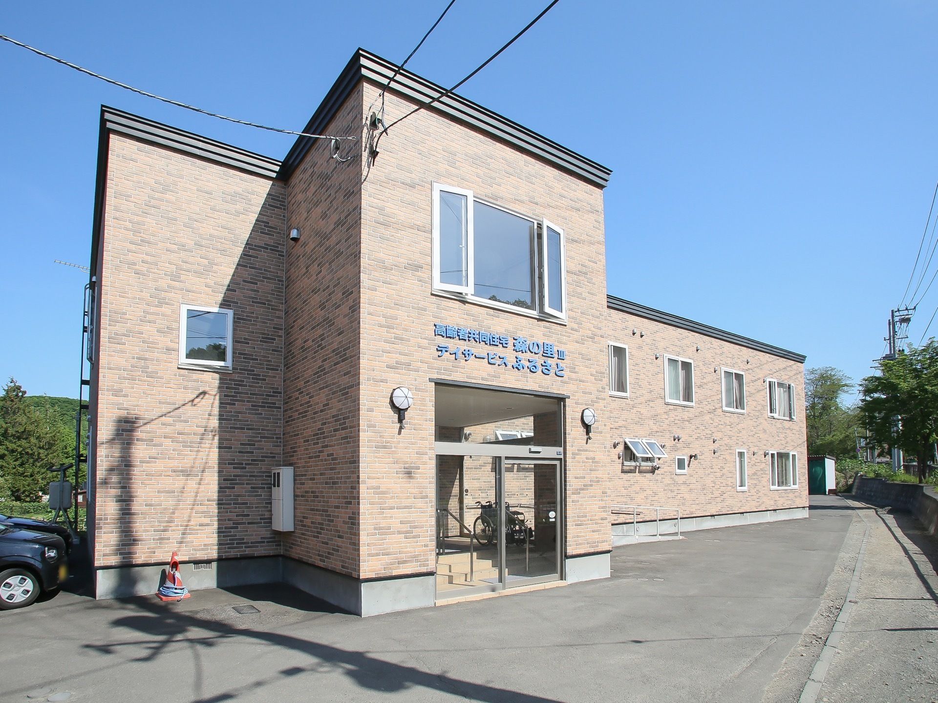 施設は茶色の外壁の２階建ての建物である。玄関上の外壁に高齢者住宅とデイサービスである旨の表示がされている。