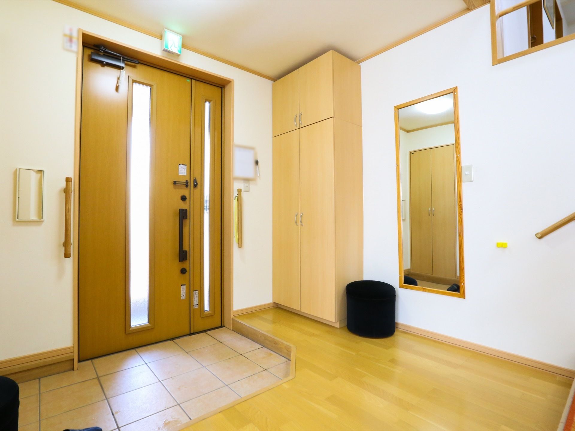 広い玄関である。大きなシューズボックスや鏡があり、黒いスツールが２つ置かれている。玄関扉の横には手すりが備わっている。