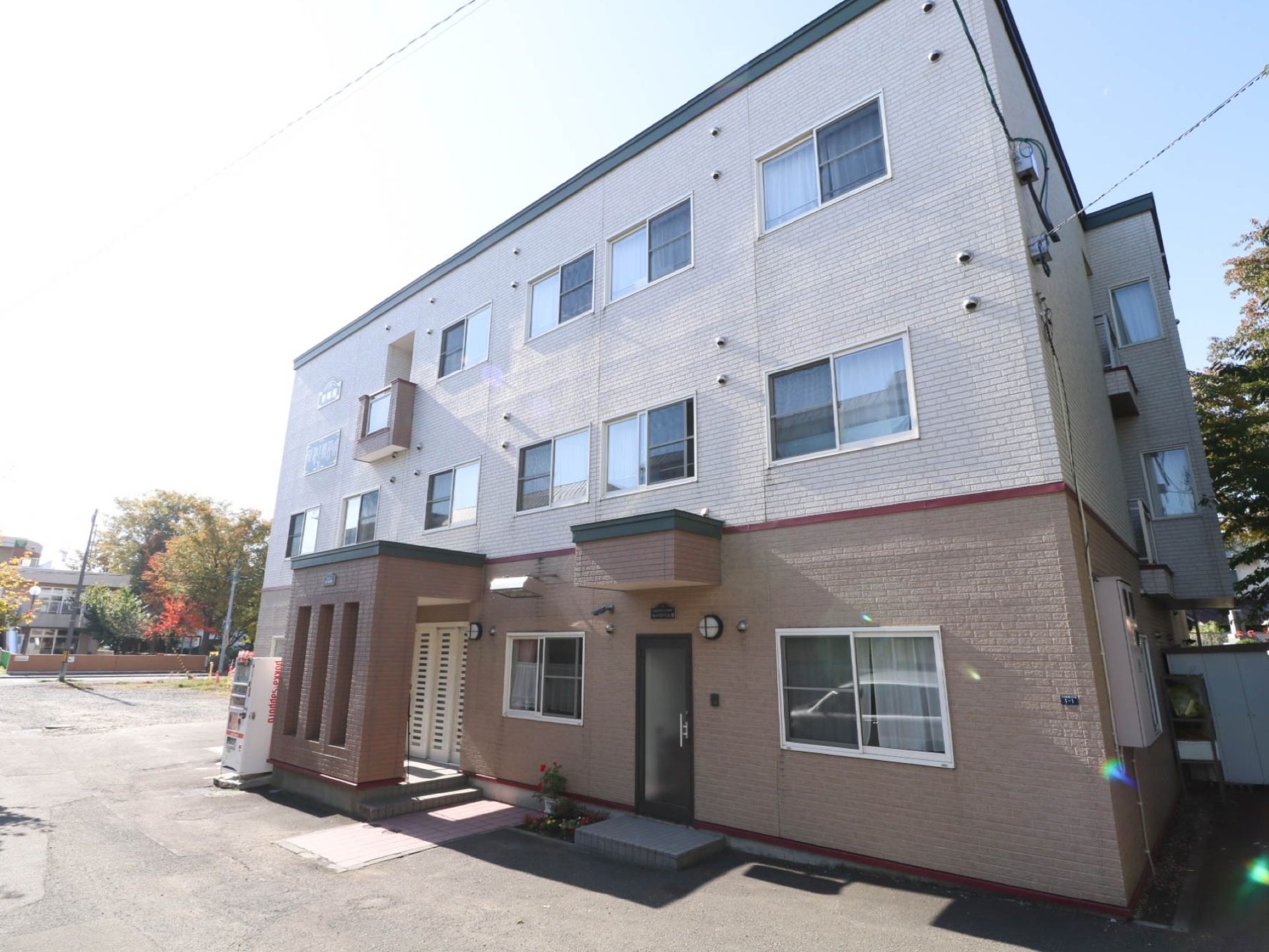 白とベージュのツートンカラーで施された施設外観である。3階建ての建物である。自動販売機が設置されている。