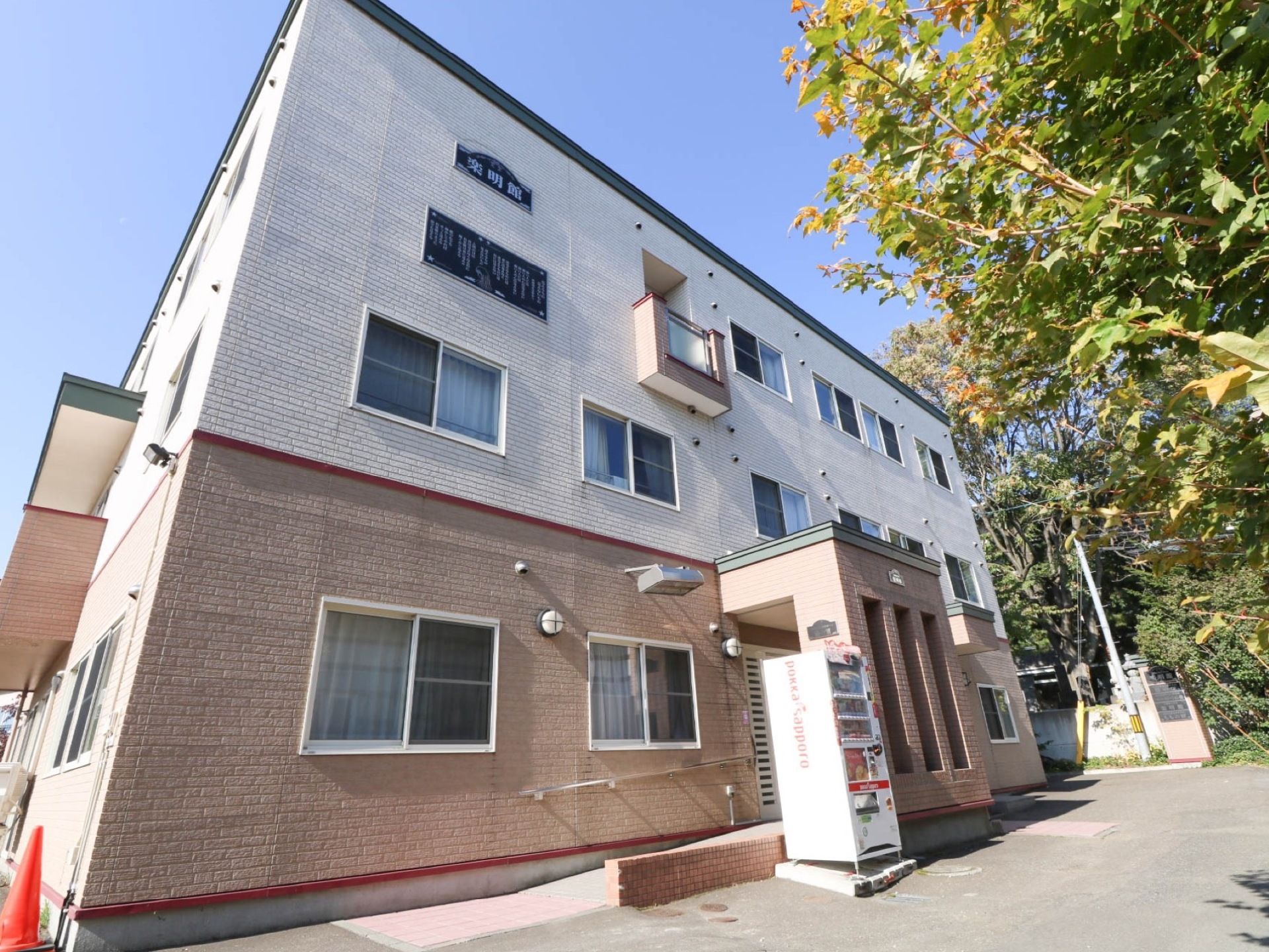 白とベージュのオシャレなツートンカラーの施設外観である。3階建て建物になっており、外に自動販売機が設置されている。