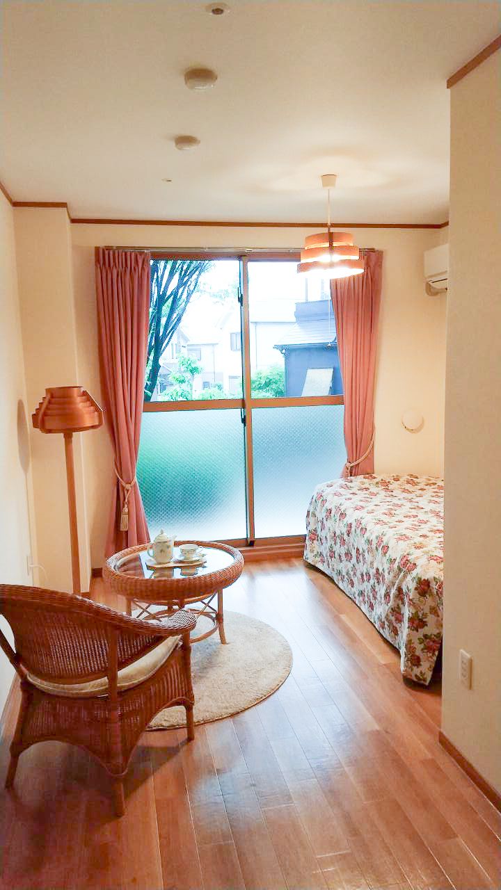 掃き出し窓から陽光がたくさん入る、明るい居室である。広いワンルームタイプで、ベッドやテーブルセット、飾り棚などが置かれている。