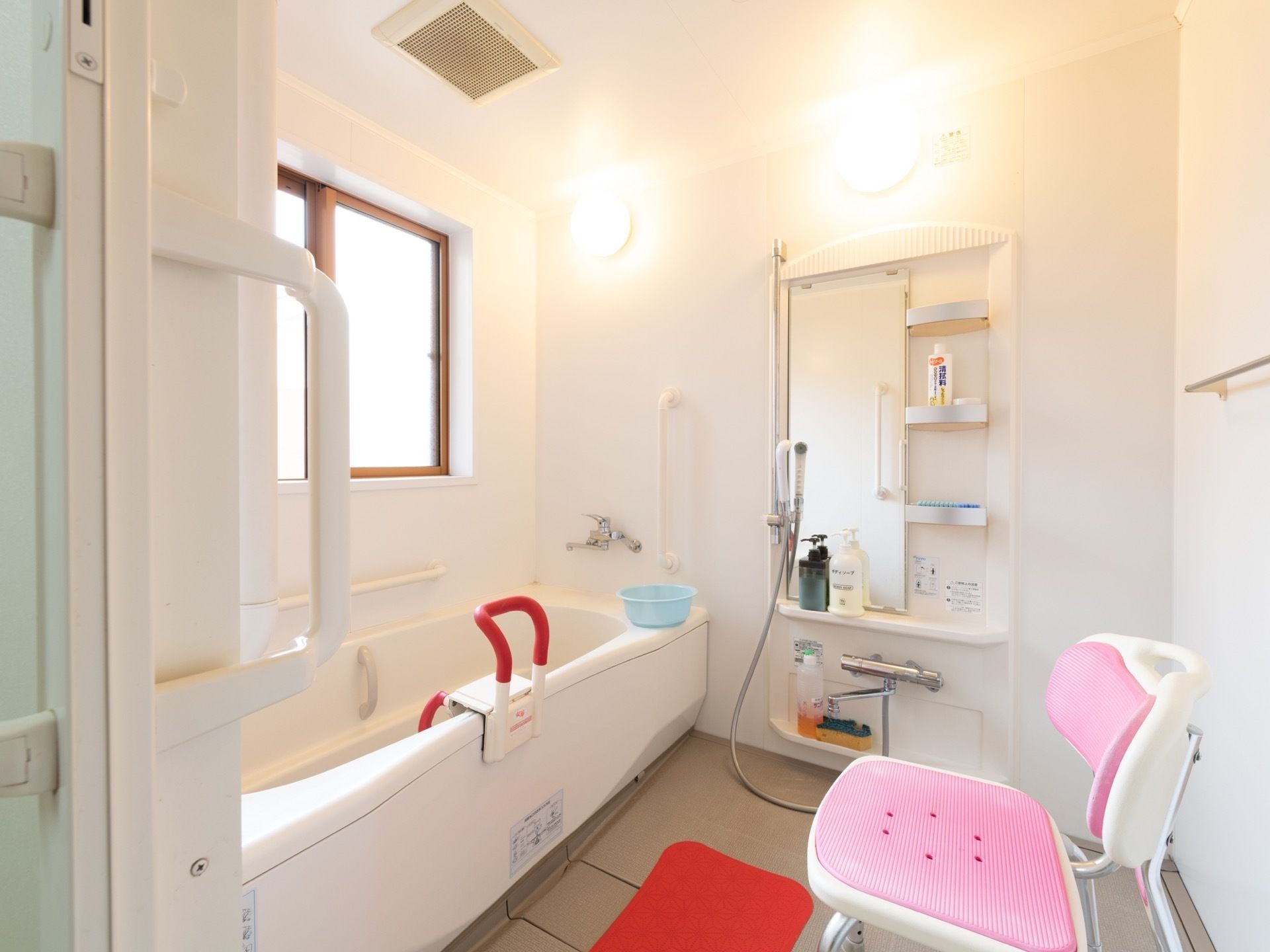 清潔感のある、広々とした白ベースの浴室である。鏡付きのシャワーにシャワーチェア、マット、手すりが設けられている。