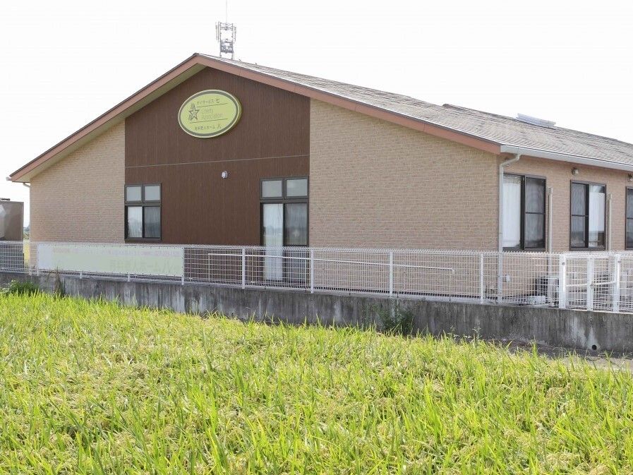 三角屋根が付いた平屋建ての建物はブラウン系で白いフェンスに囲まれ、周囲には稲が実った田んぼが広がっている。