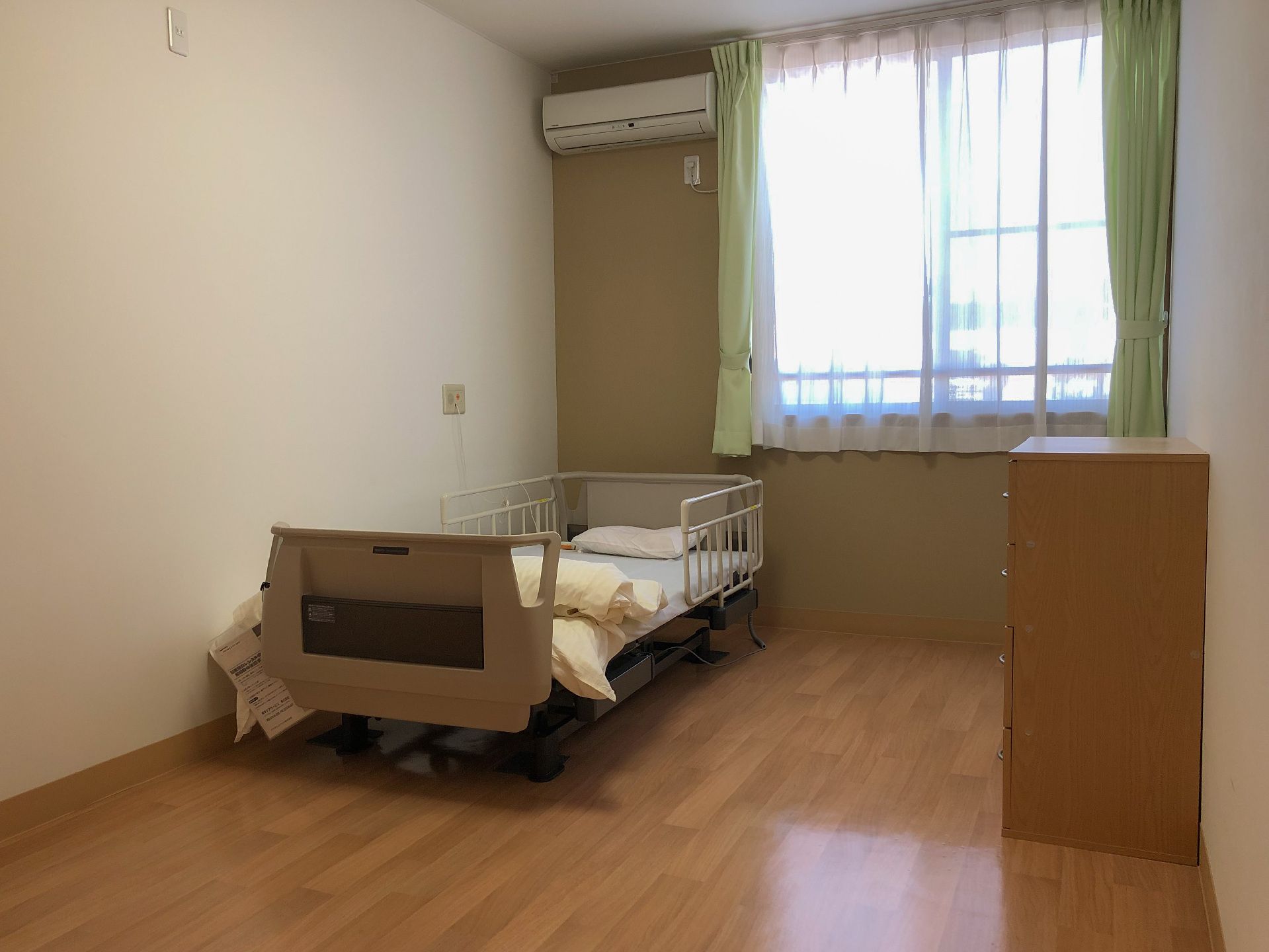 フローリングの部屋には寝具付きのベッドがあり、ベッドサイドには緊急通報装置を設置し、窓にはカーテンが付いている。