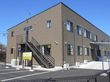 ブラウン系の２階建ての建物は玄関には屋根があり、側面には屋外階段と看板も付いている。