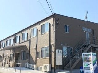 ブラウン系の２階建ての建物は玄関には屋根があり、手すり付きのスロープが設置され、自動販売機も置かれている。