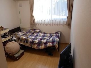 レースカーテンとカーテンが付いた大きな窓があるフローリングの部屋には寝具付きのベッドやテレビがある。