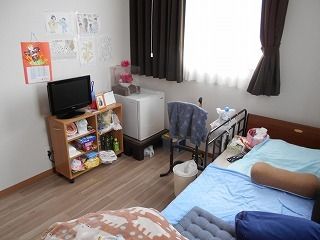 レースカーテンとカーテンが付いた大きな窓があるフローリングの部屋には、寝具付きのベッドやテレビなどもある。