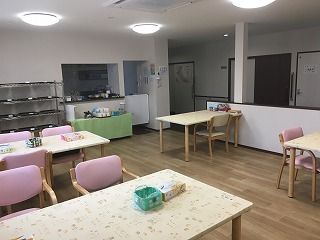 ピンクの椅子がセットされた木製のダイニングテーブルがセットされた食堂。奥には厨房があり、トレーを並べるラックもある。